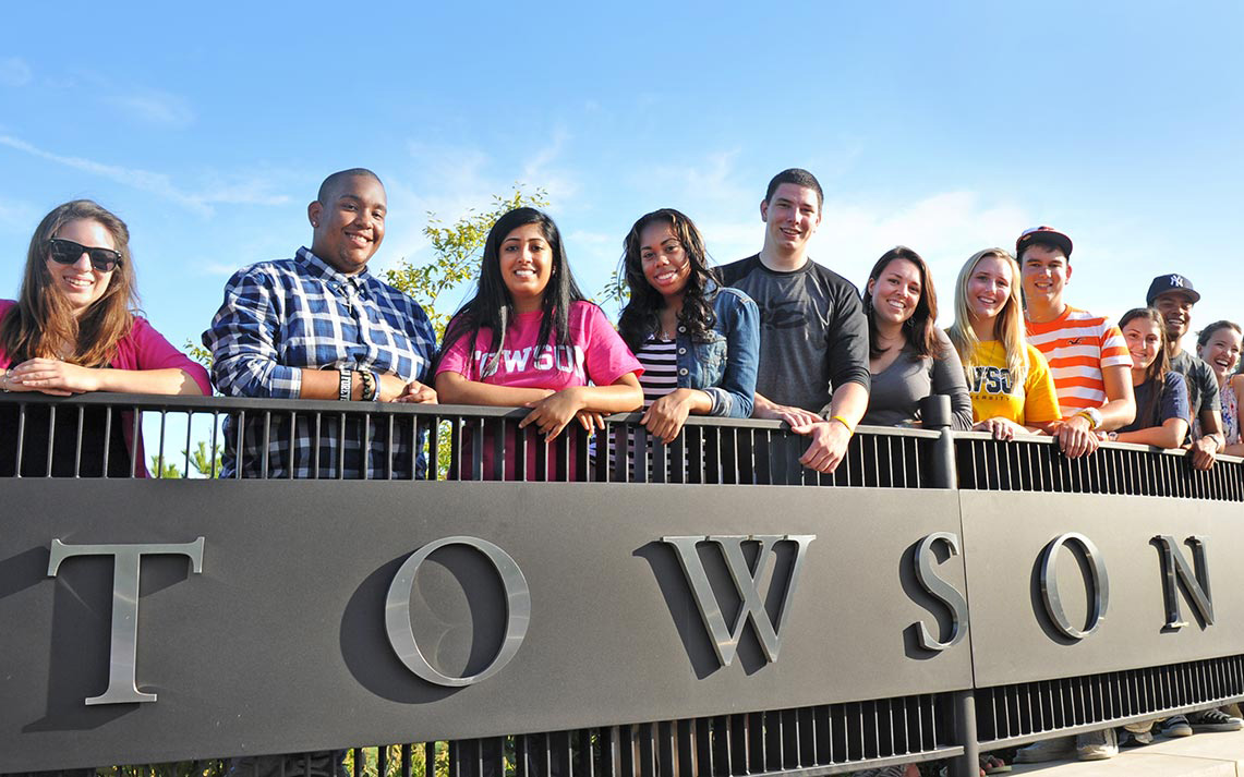 Diverse population of Towson University students standing at TU entrance sign