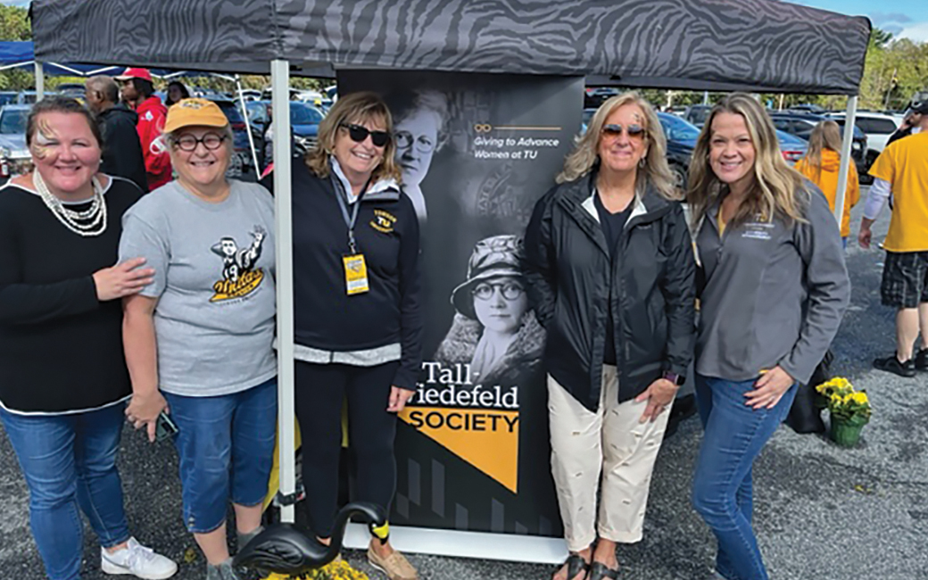 Group of women from the Tall-Wiedefeld Society stand together in front of a sign about the society.