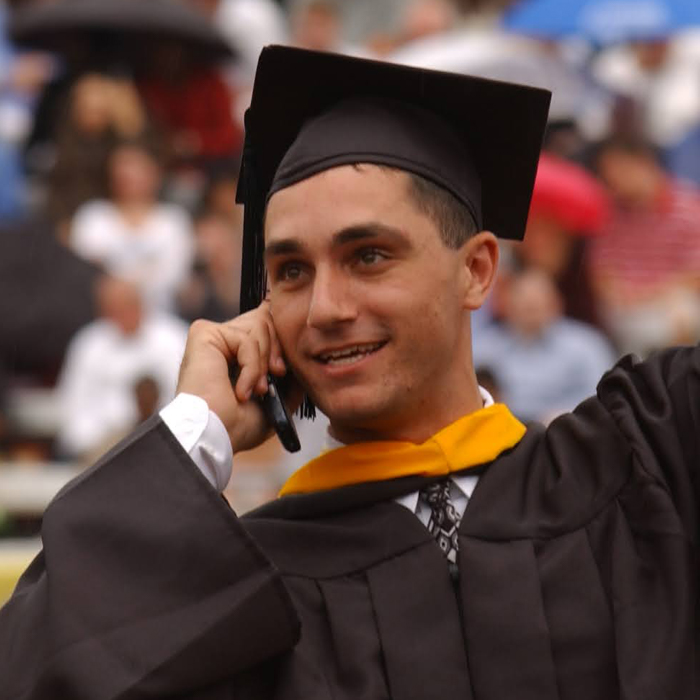 A 2005 graduate holding a flip phone and waving