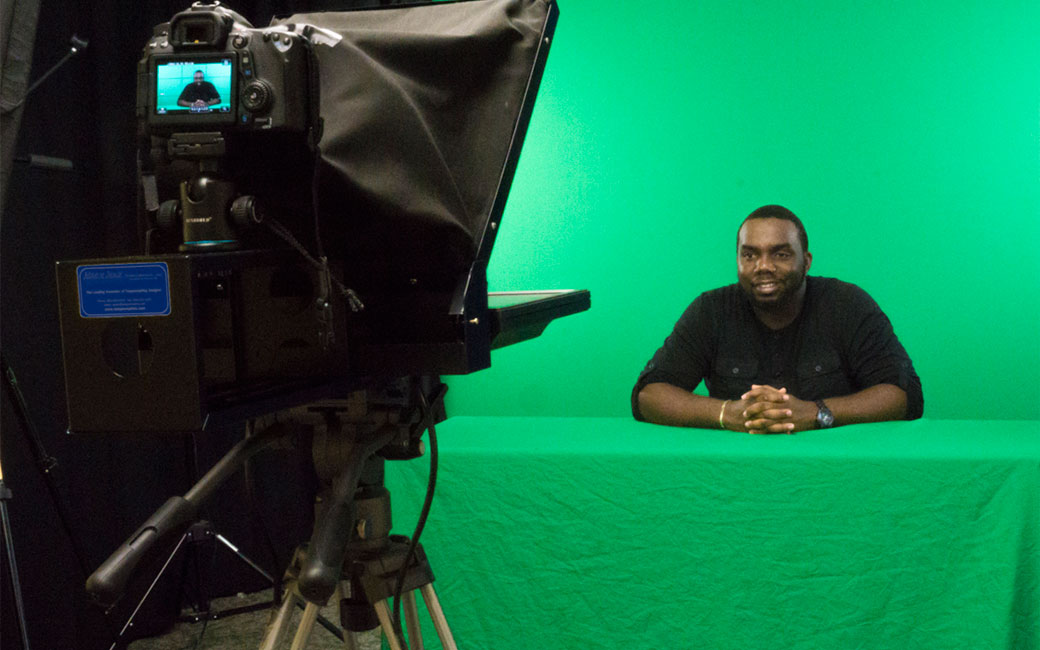 A student in SCS studio C is seated in front of a teleprompter with a green screen behind 