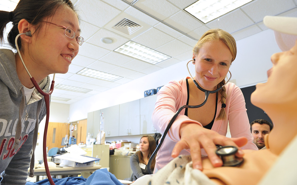 Nursing student using a stethoscope on a manikin while another student observes