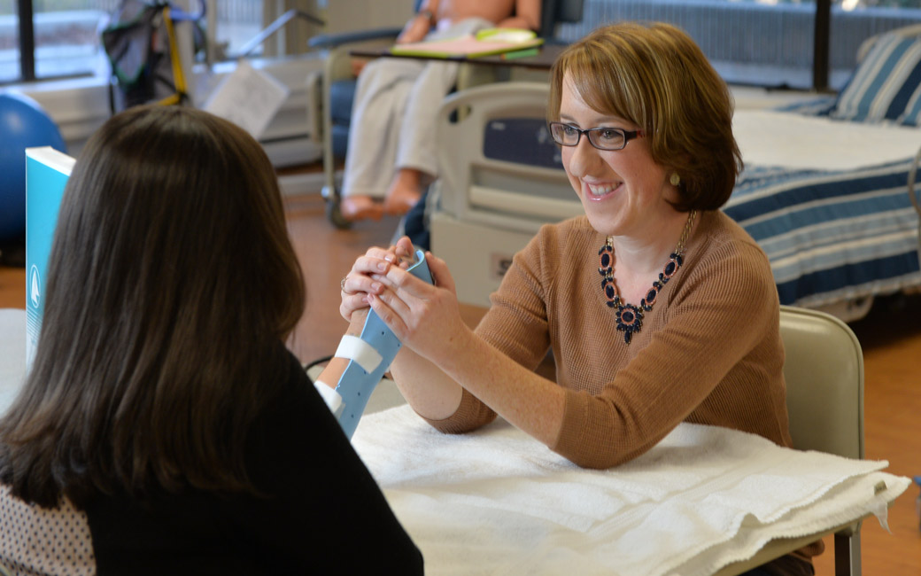 A woman in a brown three-quarter length sweater, with short brown hair and glasses, smiles as she form a blue hand splint to the patient sitting across from her. The patient has long, dark hair and a dark shirt, and sits with their back to the camera.