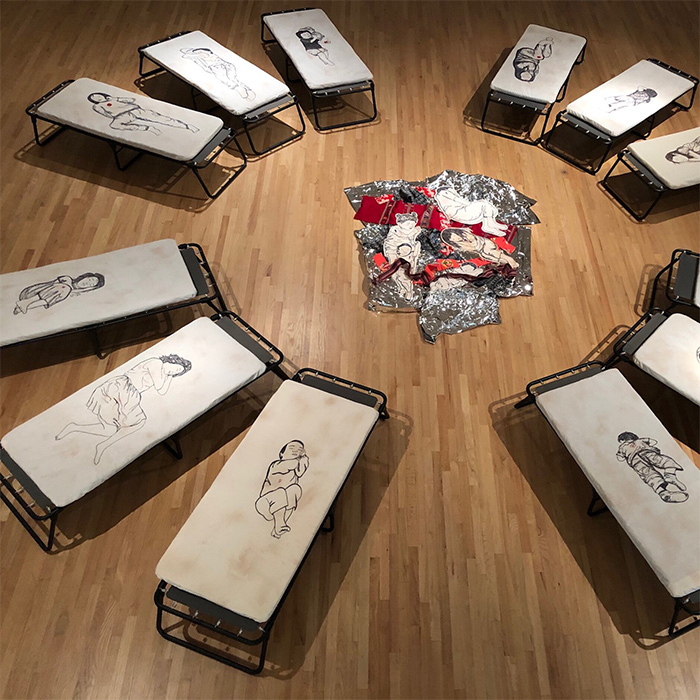 Cots sit around in a circle with drawing on them in the Dream Refuge art installation