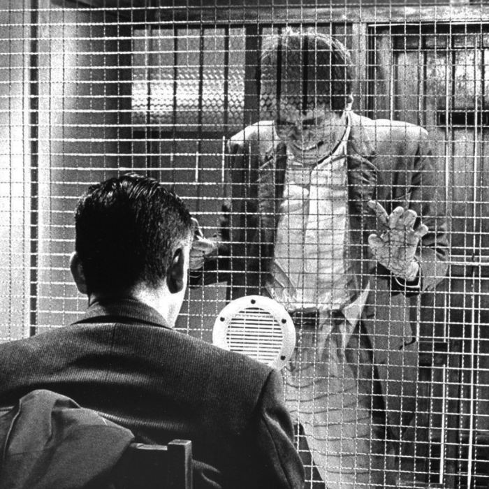 High and Low (天国と地獄, Tengoku to Jigoku) A black-and-white still from the 1963 Japanese film High and Low (directed by Akira Kurosawa), showing a dramatic prison visitation scene between two men separated by a wire mesh security screen.In the foreground, the back of a man's head and shoulders are visible as he sits facing the partition. On the opposite side of the fence, another man stands with his hands pressed flat against the wire grid. He wears a button-down shirt under a suit jacket, his expression intense and visibly strained. A circular speaker grill is mounted on the wire partition directly between the two men to facilitate communication.