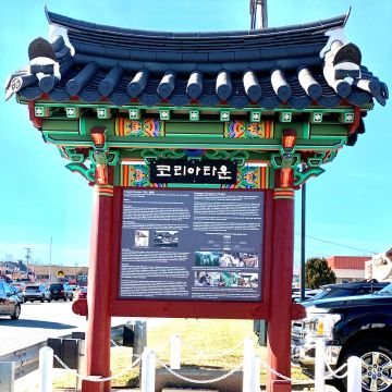 Koreatown gate A large outdoor information sign in Maryland's Koreatown featuring traditional Korean architecture. The sign is built like a gate with a dark tiled roof, colorful green and red painted wood brackets, and red columns. A black sign at the top reads '코리아타운' (Koreatown) in white Korean script. The main display board contains English text, diagrams, and historical photos under the title 'Maryland Koreatown Pavilion'.