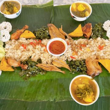 Makan Pakai Tangan A high-angle shot of a traditional Indonesian Nasi Liwet feast served on large, green banana leaves. A long bed of seasoned rice runs through the center, topped with pieces of fried chicken, fried fish, tofu, tempeh, and green leafy vegetables. A small white bowl filled with bright red sambal chili paste sits in the middle of the rice. Three small white ceramic bowls containing vegetable soup are placed around the edges, and white kerupuk crackers flank the top corners.