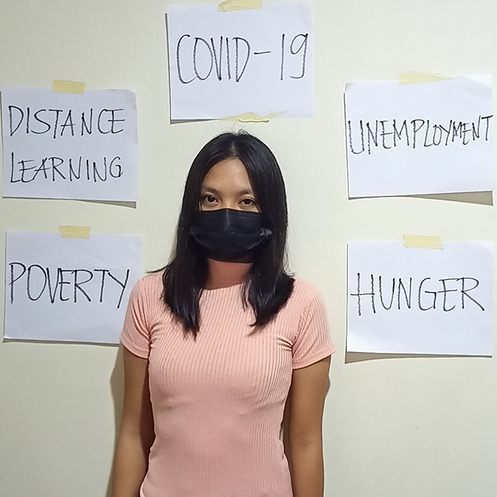 Our Stories Virtual Festival - Photo of storyteller Aya, a Filipino woman wearing a black mask and pink short sleeve short. Taped on the wall behind her are the words "POVERTY," "DISTANCE LEARNING," "COVID-19,"UNEMPLOYMENT" and "HUNGER."