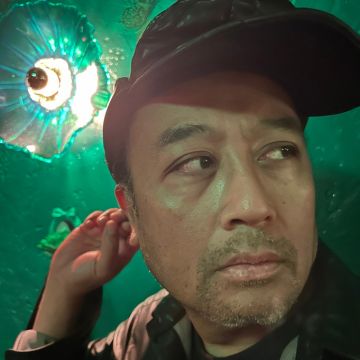 Regie Cabico A close-up portrait of Regie Cabico looking off-camera to his right under dramatic green lighting. He is wearing a dark baseball cap and has slight stubble on his face. He raises his right hand toward his ear. In the background, a bright, circular light fixture shines through a textured, translucent green glass surface, creating a vibrant aquatic or atmospheric glow.