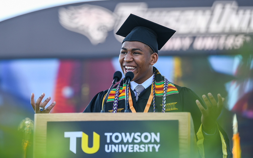 Student in commencement regalia smiling while delivering speech from stage