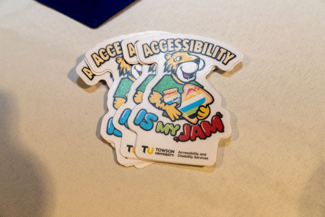 A pack of stickers feature the Towson University mascot, Doc, a black and gold cartoon tiger, holding a piece of toast with the disability pride flag spread on top of it. The sticker reads, "Accessibility is my jam".