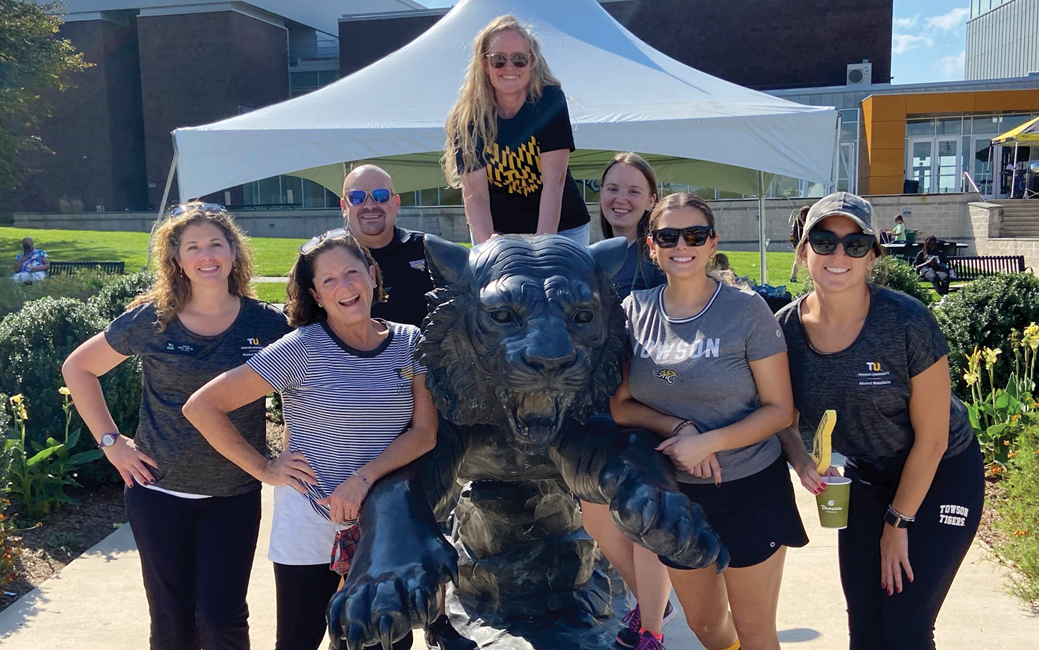 A group of seven people, one perched on a Tiger statue, smiling outside SECU Arena