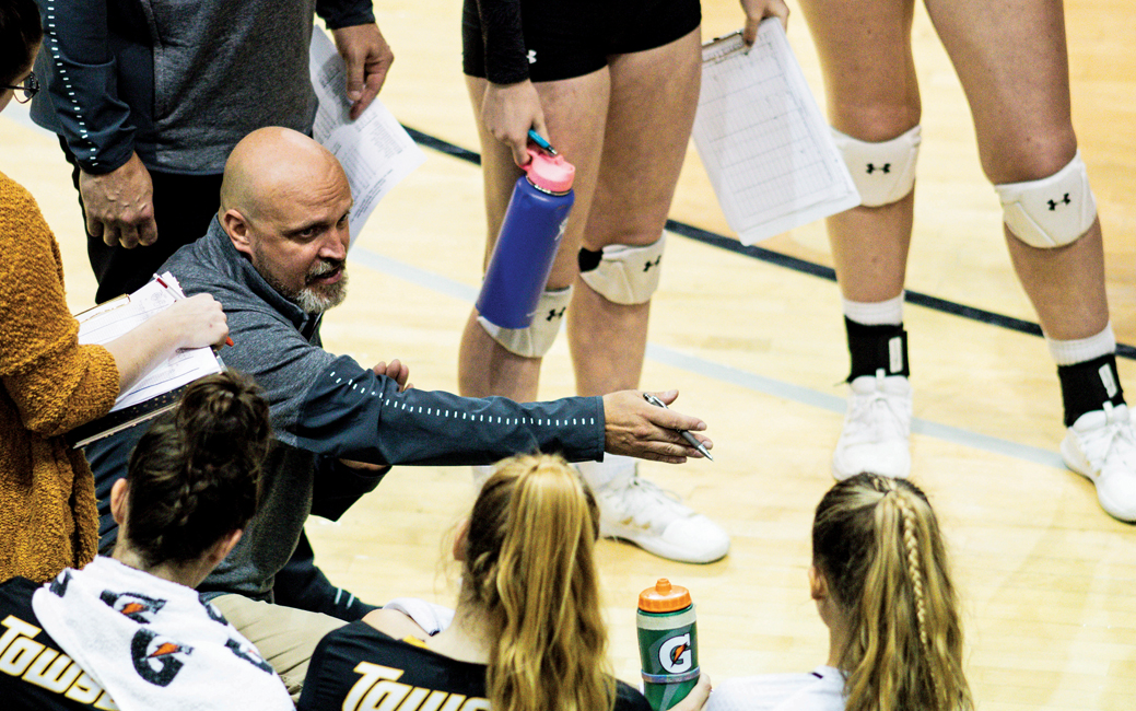 Photo of a Don Metil coaching a women's volleyball team during a timeout, gesturing