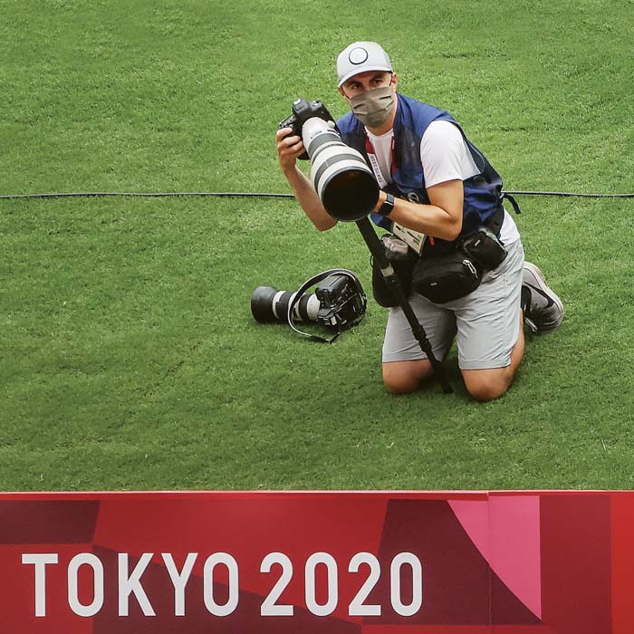 Patrick Smith kneeling on grass holding a camera in front of a red banner that says Tokyo Olympics