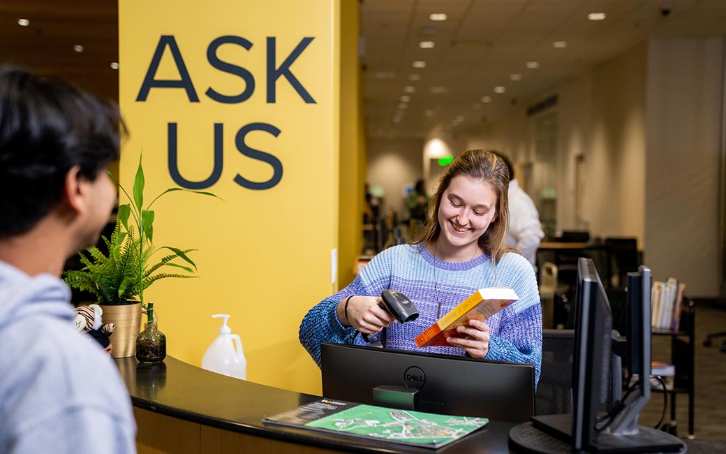 Student scans book at counter with words "Ask Us" in background