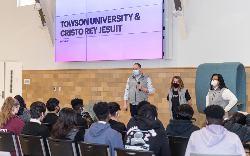 Speakers in front of large screen reading "Towson University & Cristo Rey Jesuit" and audience