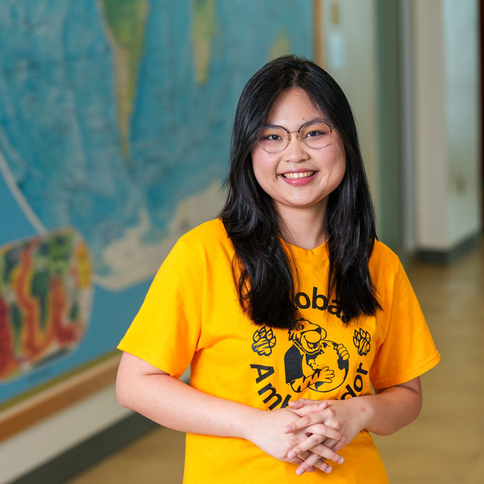 Sandy smiles, hands clasped in front of one of the several world maps in the Liberal Arts Building