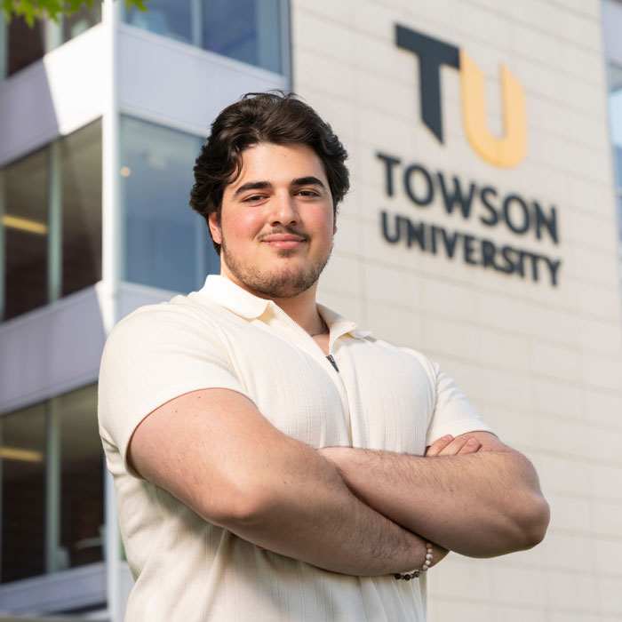 A scruffy male student stands crossarmed before a building with Towson University branding