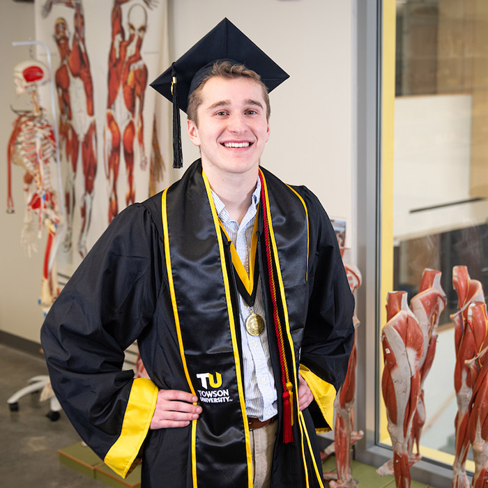 Graduate in cap and gown poses in classroom with anatomical models in background