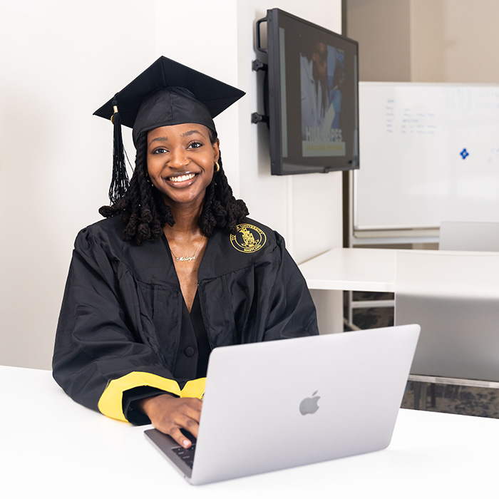 student in cap and gown sitting with laptop