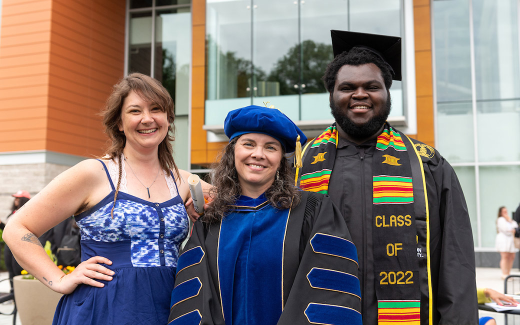 three people pose, two wearing graduation regalia