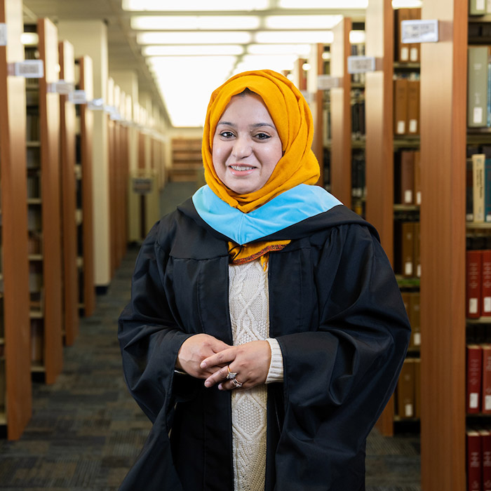 Graduate poses in library wearing cap, gown and blue master's hood