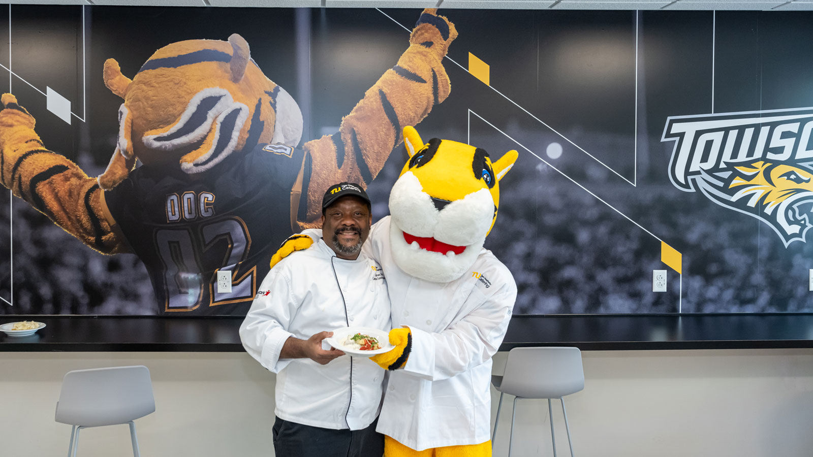 TU Chef and Doc stand next to a mural in Doc's South Campus Kitchen