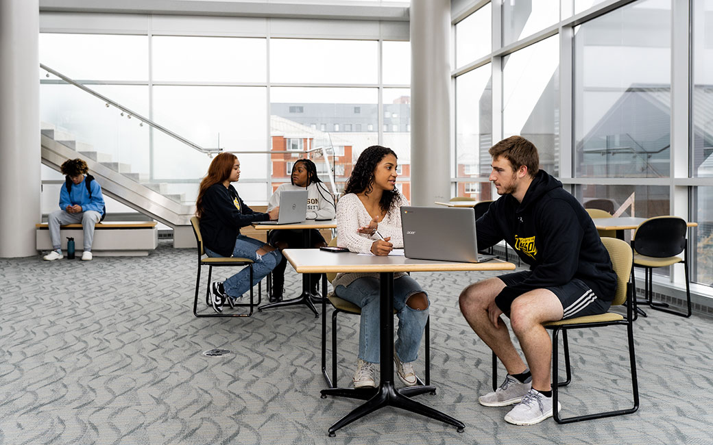 Students sit at table looking at laptop in study area