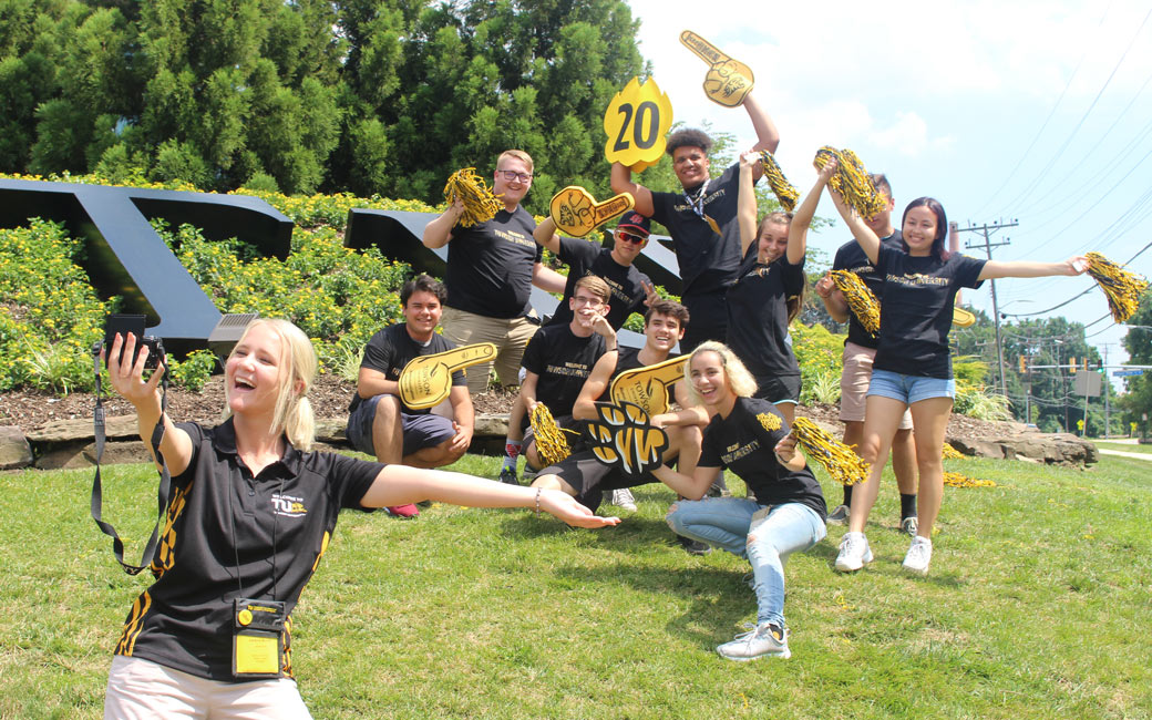 Student leader taking a selfie with a group of incoming students in TU shirts by a TU logo statue